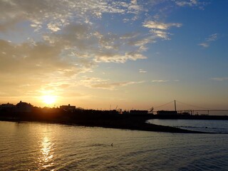 Naklejka premium 白い雲が浮かぶ青空に上ってくる朝日とその光が映りオレンジに輝く河口の水面