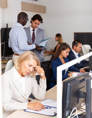 Upset and tired elderly woman sitting at desk in open plan office with busy colleagues on background
