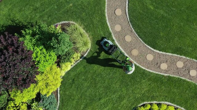 Professional Male Gardener Mowing Large Area Of Green Lawn On Well Kept Property. Aerial View Of Beautifully Designed Garden With Variety Of Plants. 