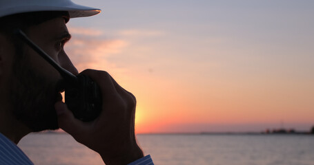 Close-up of an engineer talking into a walkie-talkie and looking towards the sea at dawn. - obrazy, fototapety, plakaty