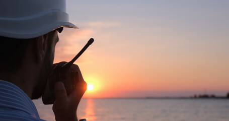 Close-up of an engineer talking into a walkie-talkie and looking towards the sea at dawn.