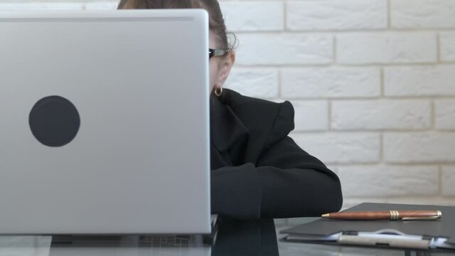 Curious Child Type On Computer. A Little Happy Girl Looks Out From Behind The Screen And Shows Her Tongue In The Office.