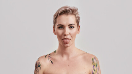 Portrait of a young attractive half naked tattooed woman with perfect skin showing her tongue to camera isolated over light background