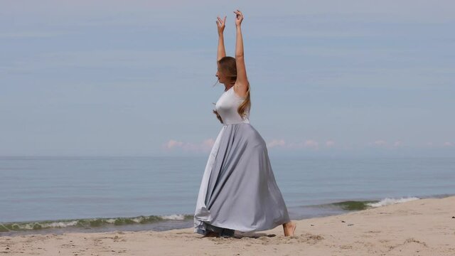 Beautiful young woman in a long dress dancing on the beach against the blue sky on a sunny summer day
