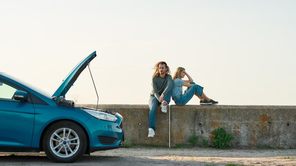 Outdoor shot of couple of travelers sitting near the broken car with open hood, Man and woman...