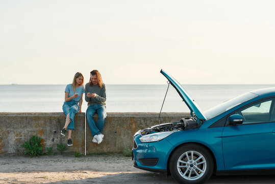 Outdoor shot of couple of travelers having problems with the car, man and woman sitting near broken car with open hood and looking for a phone number of assistance, trying to solve problem - Powered by Adobe