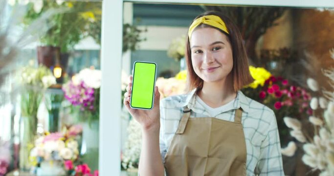 Charming young woman florist wearing apron in modern flower store. Attractive Caucasian female worker holding green screen, chroma key, smiling and looking to camera. Business, commerce concept.