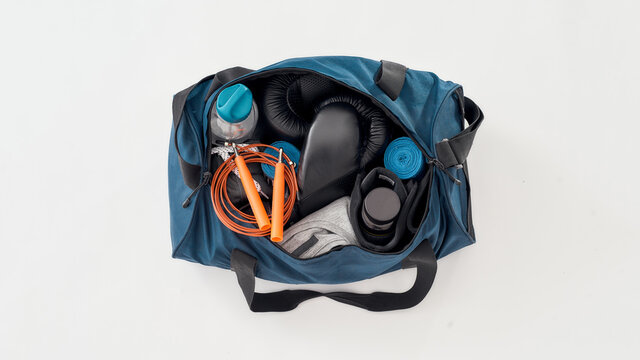 Top view of a sports bag with gym equipment isolated on grey background. Boxing gloves, hand wraps, clothing, bottle of water and jumping rope