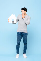 Full length portrait of thoughtful Caucasian man holding house model cutout and thinking in light blue isolated studio background
