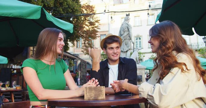 Portrait of happy Caucasian friends sitting at table outdoors and chatting. Handsome boy in hat and beautiful girls smiling and talking at cafeteria terrace in town in good mood. Friendship concept