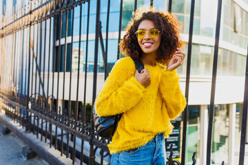 Outdoor bright portrait of happy African girl with back pack  and standing on urban background. Wearing yellow sweater and sunglasses. Pretty student enjoying sunny spring day.