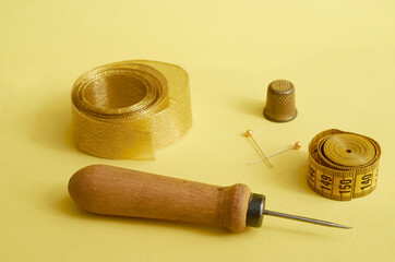 Sewing supplies. Awl, gold ribbon, sartorial meter, thimble and two pins on a yellow background
