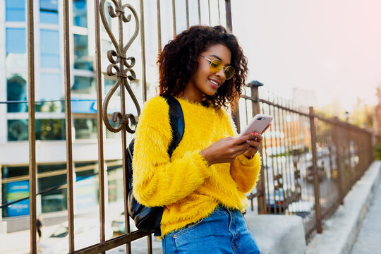 Outdoor image of young attractive black woman with stylish Afro hairs  using mobile phone and sitting on  bridge  in modern district. Yellow glasses and pullover.