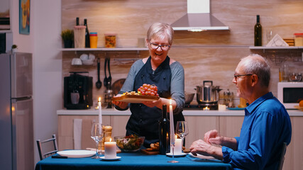 Old senior woman serving her husband with grapes and cheese. Elderly old couple talking, sitting at the table in kitchen, enjoying the meal, celebrating their anniversary at home with healty food.