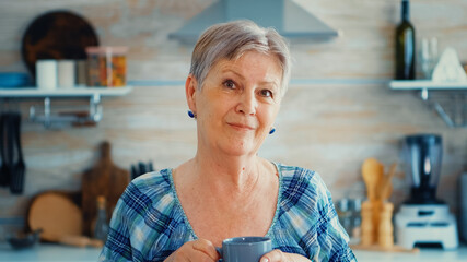Winkled senior woman smiling at camera holding cup of offee in kitchen during breakfast. Authentic...