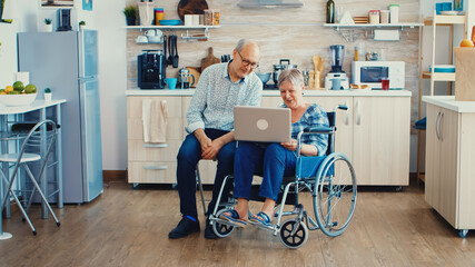 Senior couple waving at webcam during a video call on laptop in kitchen. Paralysied handicapped old elderly woman and her husband on online call, using modern communication tech.