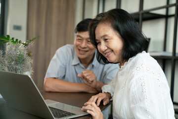 Happy retirement life and insurance concept.Senior asian couple sitting on sofa in home playing laptop and relaxing together. They are smiling to spend their time together with happiness at home.