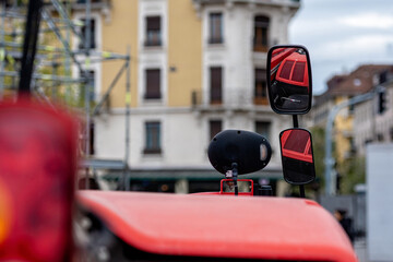 Close-up of tractor side mirror on defocused city background. The mirror shows the red hood of the tractor.