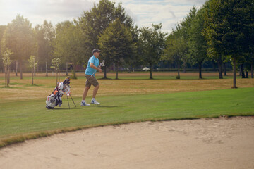 Man playing golf going to the green with his putter that he has just taken from his golf bag with a sand hazard or bunker in the foreground