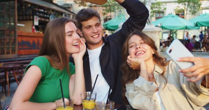 Portrait of joyful Caucasian handsome guy and pretty girls making selfie photos on smartphone while sitting at table outdoor. Happy friends taking pictures on cellphone in cafeteria. Leisure concept