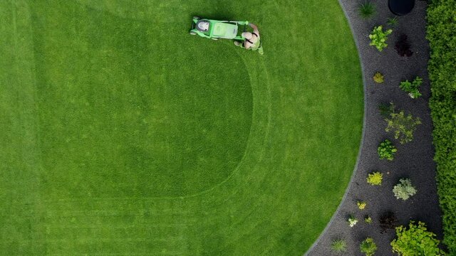Aerial View Of Male Gardener With Push Mower Neatly Cutting Green Grass In Circular Pattern. Lawn Maintenance. 