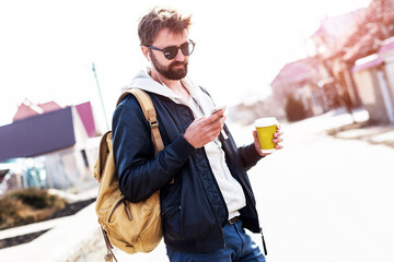 Happy hipster man with beard and sunglasses typing message and walking in the countryside, drink coffee and listen Music. Wearing stylish leather jacket, backpack and hoodie.