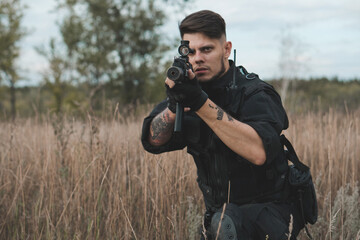 Young soldier in black uniform sitting and aiming with an assault rifle.