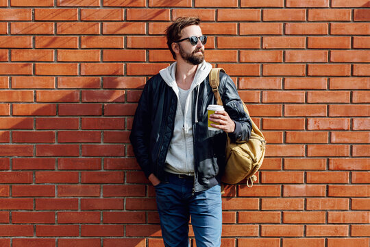 Outdoor fall or winter portrait of handsome hipster man with beard in hat , grey hoodie and leather jacket with bag pack holding cup of hot coffee. Brick urban wall background.