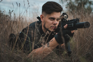 Young soldier in black uniform lying down and aiming an assault rifle.