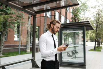 A curly-haired guy, in a white shirt and tie, at the bus stop looks at the phone while waiting for the bus. The concept of a mobile application for traffic in the city.