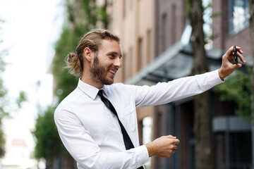Portrait of a successful businessman taking a selfie and smiling. A curly-haired manager, in a white shirt and tie, happily takes a selfie on a phone camera.
