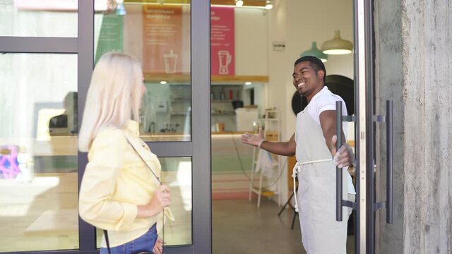 Good-looking smiling man in uniform welcomes customers in their shop, glad to see blonde caucasian lady.