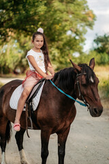 Fototapeta premium Little blonde girl with long hair rides a horse in the park at sunset in autumn. Autumn horse ride. Friendship of a girl and a horse.