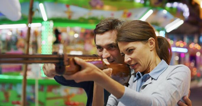 Authentic shot of a happy carefree smiling couple in love is having fun to play fair shooting games and win in amusement park with luna park lights at night.