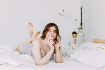Beautiful cute brunette girl in pajamas communicates, types on the phone and thinks deeply lying in...