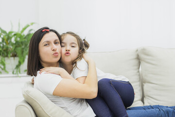 Obraz premium Photo of mother and daughter sitting on white sofa and show kiss on camera.