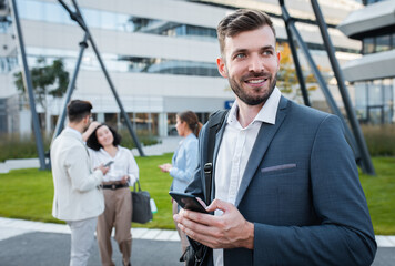 Naklejka na ściany i meble Portrait of smiling businesswoman with smartphone in his hands standing in front of modern office buildings with colleagues behind him.