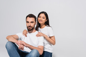 brunette asian woman touching boyfriend and looking at camera while sitting together isolated on white