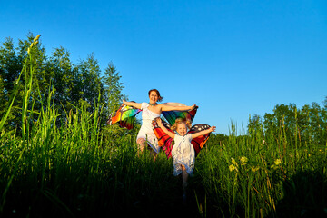 Mother and daughter with bright fabric like butterfly wings play, fly, run, jump in meadow or field with green grass and dandelion flowers. Young woman and a little girl have fun on natural landscape.