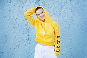 Smiling young woman with short shaved hair and piercing wears yellow hoodie against blue wall.