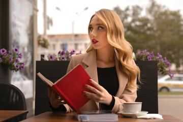 Portrait of Young modern girl, sitting at cafe outside with cup of coffee and book in the street