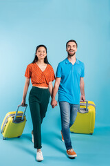 full length view of interracial couple of tourists holding hands while walking with suitcases on blue