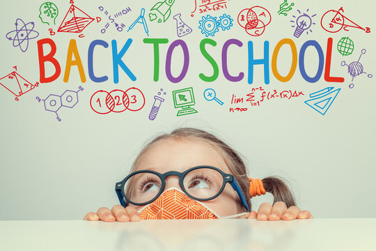 Beautiful cute little girl wearing protection mask against Covid-19 virus and hiding under table. Curious girl looking at back to school word and illustrations above her head.