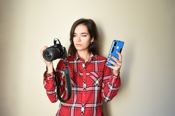 Young brunette woman photographer in red shirt choosing between camera and mobile phone on beige background. Concept of choice and difference between mobile photography and camera. Copy space