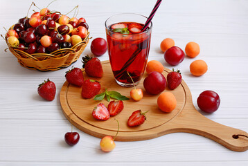 Strawberries, plums, mint leaves and a glass of juice are on the cutting board, the cherry lies in a wicker basket on a light wooden table