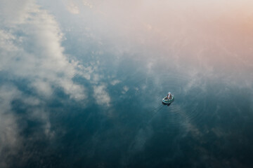 Lonely fisherman in a boat among the lake reflection of sky and clouds in smooth water. Calm and relaxation hobby man with fishing rod fishing early in the morning. Aerial view