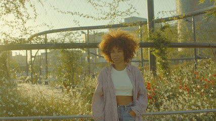 Portrait of mixed race black woman in the park full of flowers with huge afro hair in contour sunlight watching to the camera. Cinema film colors