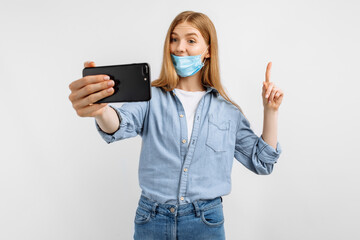 Happy young business woman in a medical protective mask on her face, talking on a mobile phone video link, on a white background