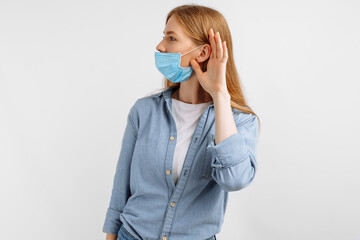curious, focused young woman in a medical protective mask on her face, listening with her hand to...