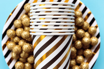 Golden, white and black striped paper glasses close up with dish on blue  background with decorative balls.